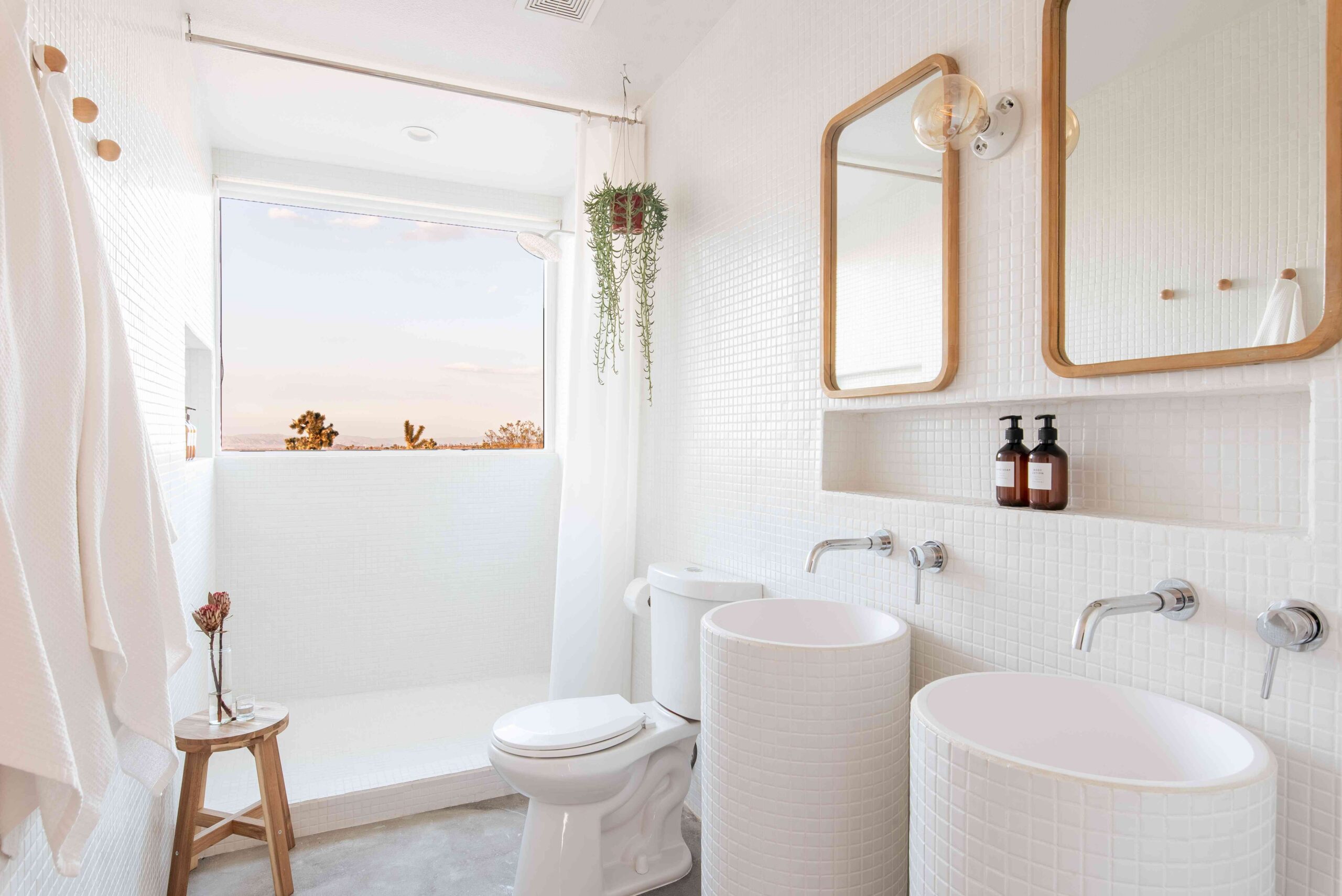 Bright minimalist bathroom with dual pedestal sinks, white mosaic tile walls, and open shower with scenic outdoor view.