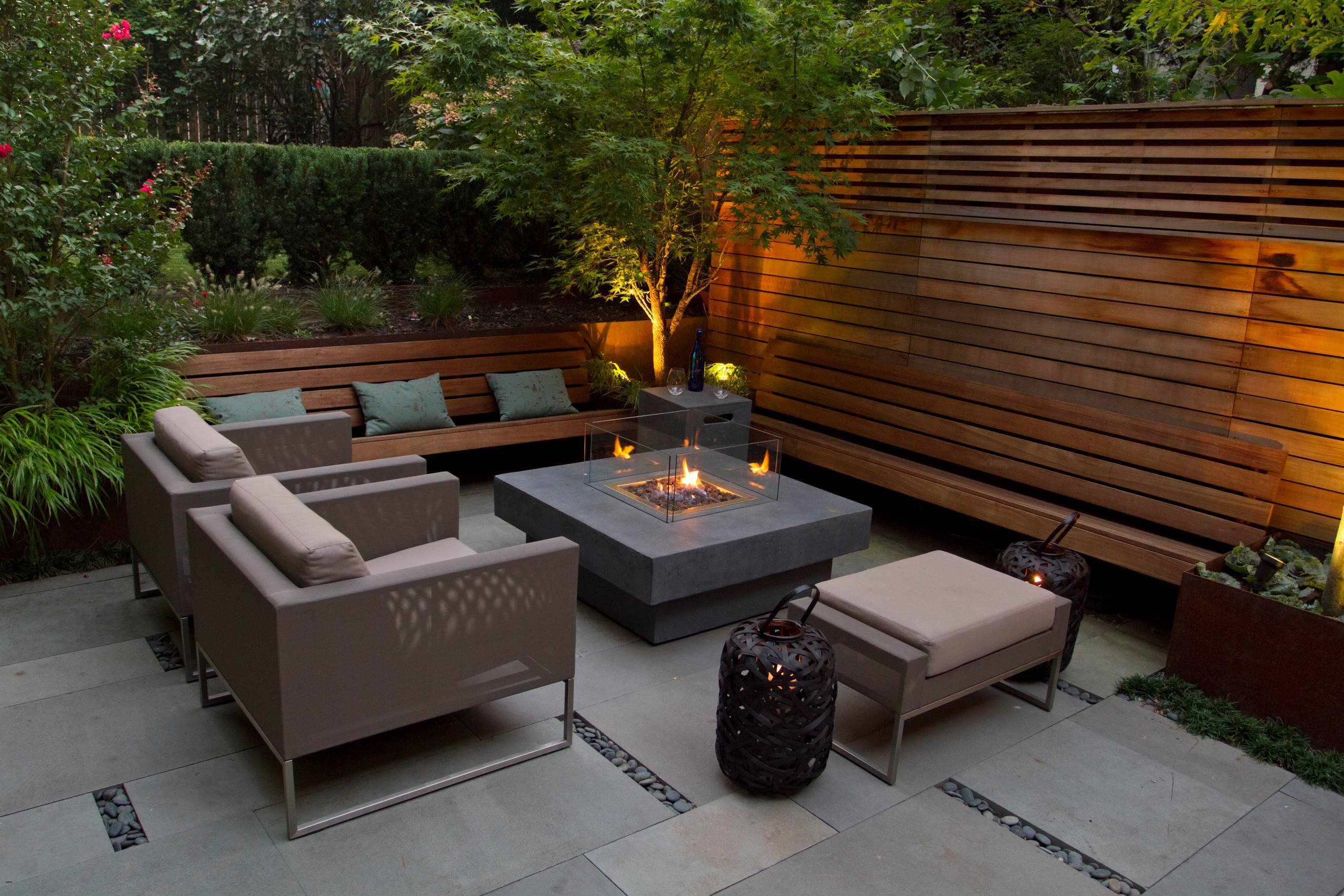 An intimate backyard seating area featuring a modern fire pit surrounded by beige armchairs and a matching ottoman. A wooden bench with green cushions lines the back, complemented by a wooden privacy fence and lush greenery.