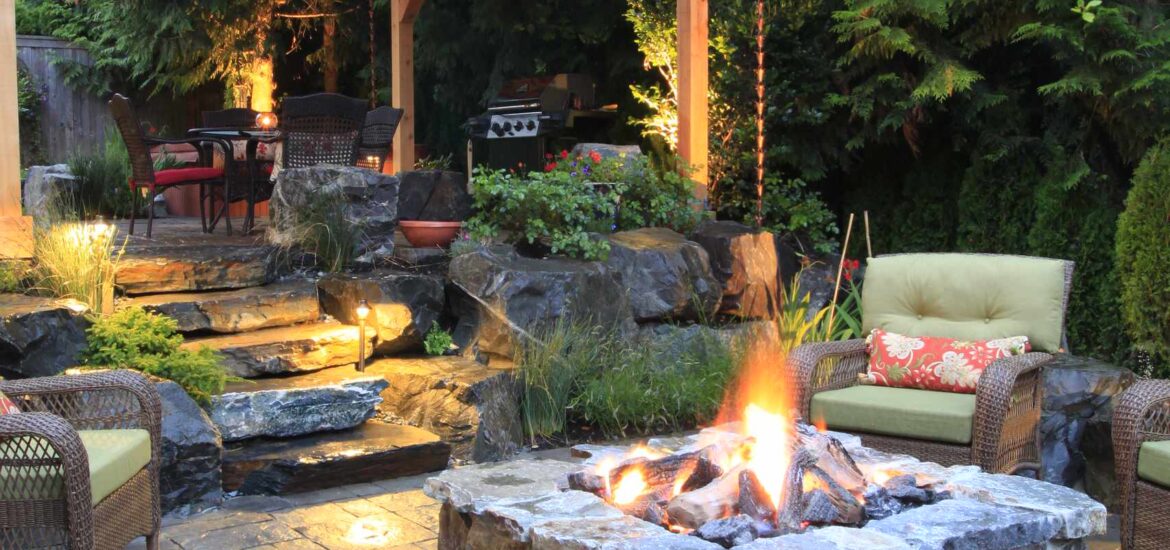 A rustic backyard patio featuring a stone fire pit surrounded by wicker armchairs with green cushions. A wooden pavilion with a pitched roof provides shelter for dining and grilling, while stone pathways and lush greenery create a natural ambiance.
