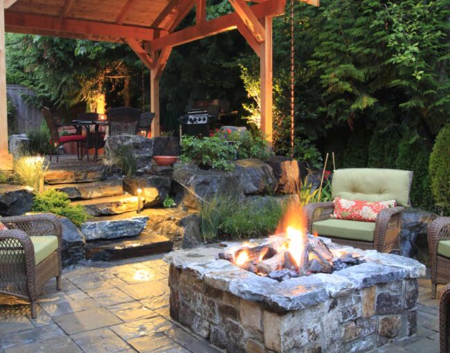 A rustic backyard patio featuring a stone fire pit surrounded by wicker armchairs with green cushions. A wooden pavilion with a pitched roof provides shelter for dining and grilling, while stone pathways and lush greenery create a natural ambiance.
