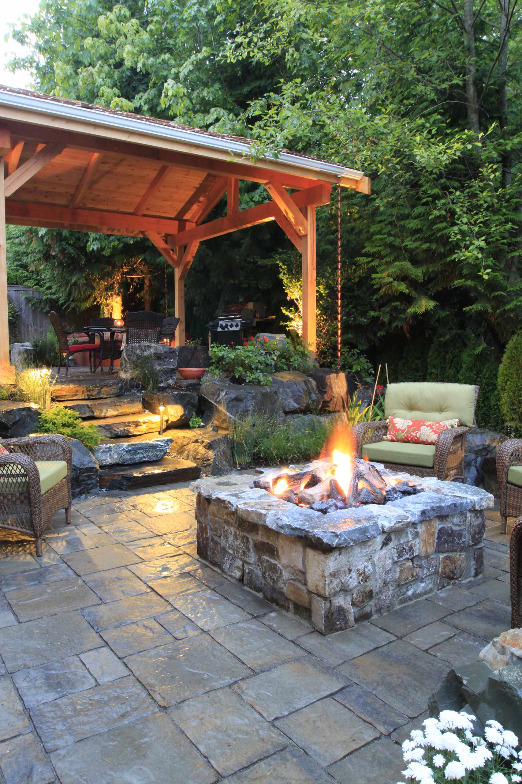 A rustic backyard patio featuring a stone fire pit surrounded by wicker armchairs with green cushions. A wooden pavilion with a pitched roof provides shelter for dining and grilling, while stone pathways and lush greenery create a natural ambiance.