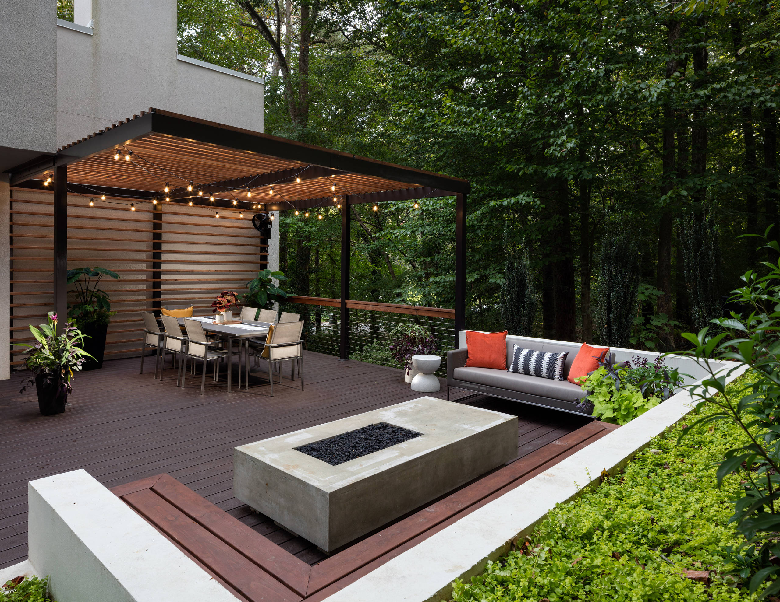 A modern backyard retreat featuring a covered dining area with string lights, a sleek concrete fire pit, and comfortable seating with orange and striped cushions. Surrounded by lush greenery and forest views, the space combines natural and contemporary elements.