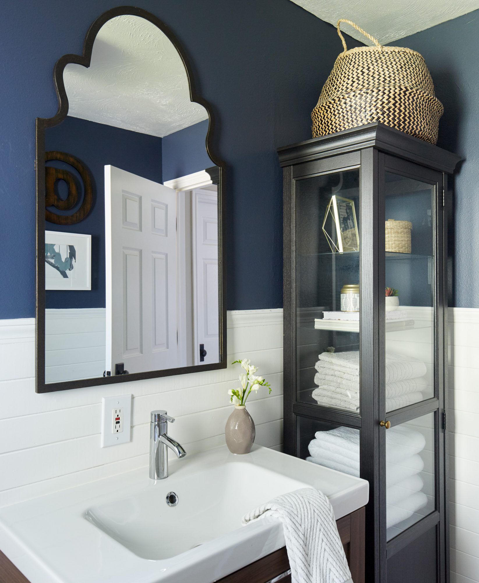 Small bathroom with navy blue walls, a white vanity, and a tall black glass cabinet for towel storage.
