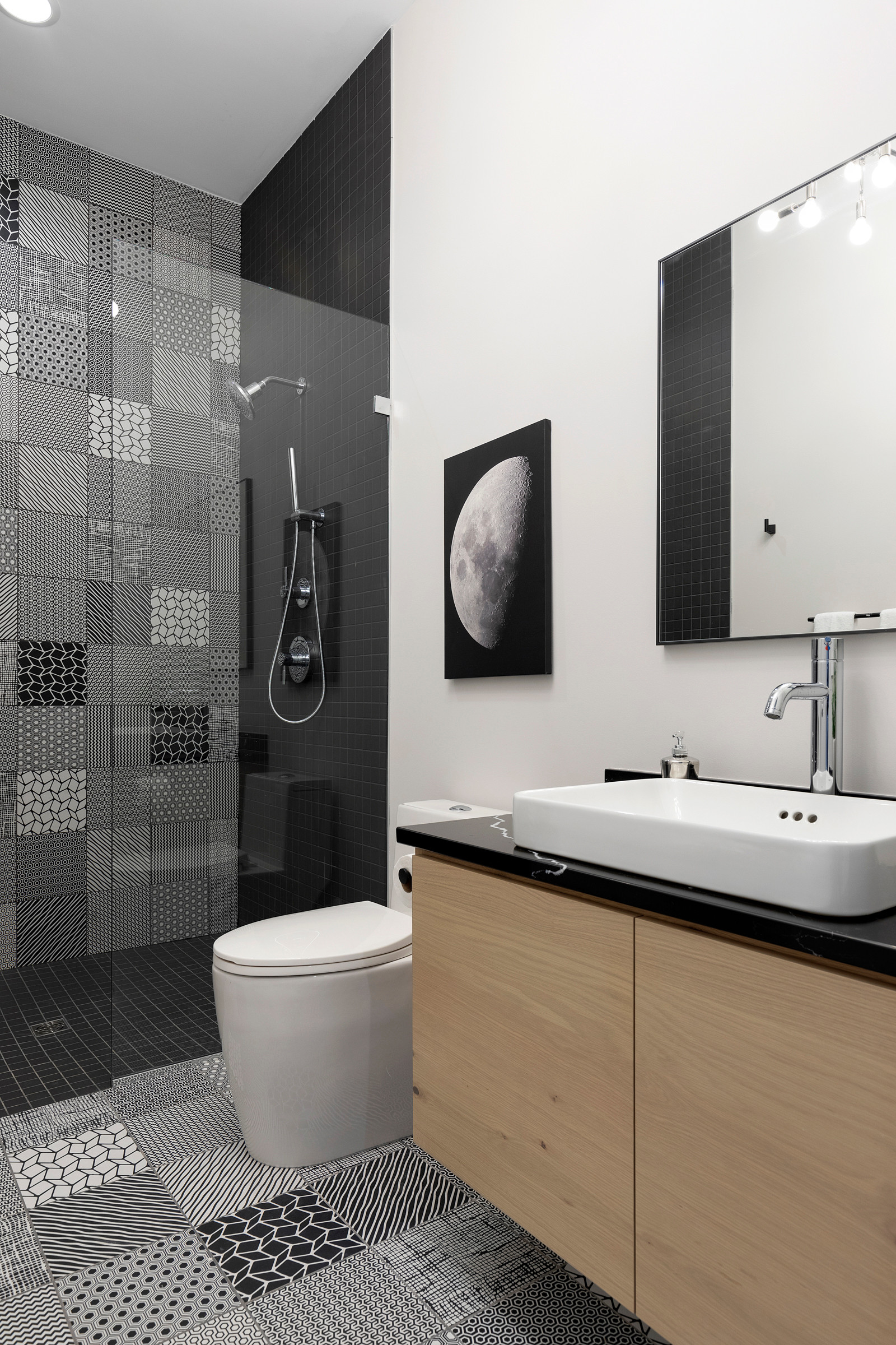 Modern bathroom with black and white patterned tiles, glass shower, and wooden vanity.