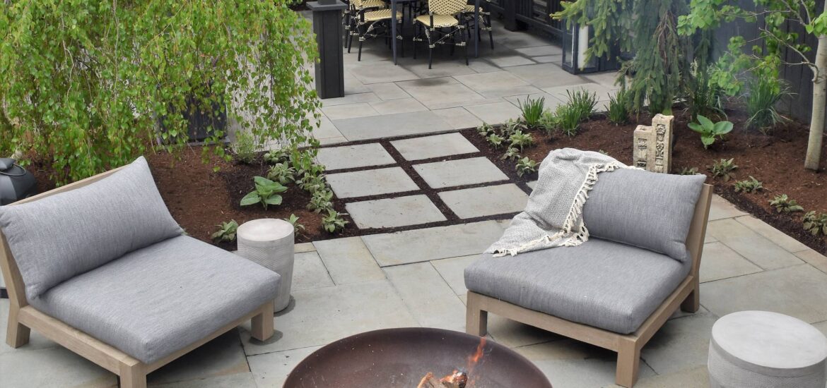 A modern backyard design featuring a cozy seating area with gray cushioned chairs surrounding a fire pit, a dining area under a black pergola, and lush greenery with trees and plants enhancing the ambiance.