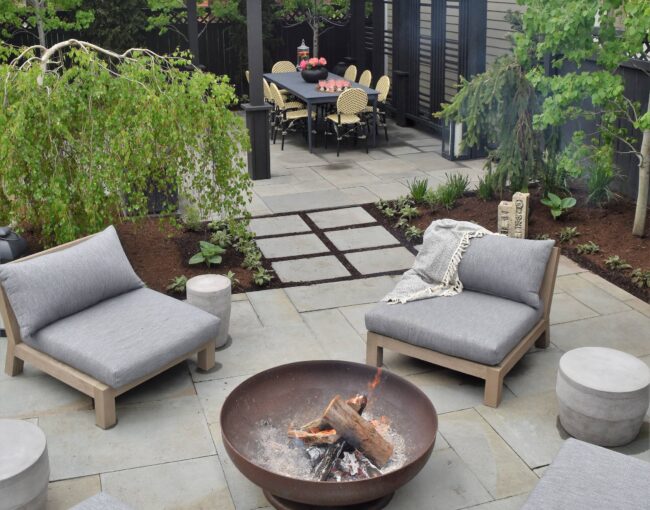 A modern backyard design featuring a cozy seating area with gray cushioned chairs surrounding a fire pit, a dining area under a black pergola, and lush greenery with trees and plants enhancing the ambiance.