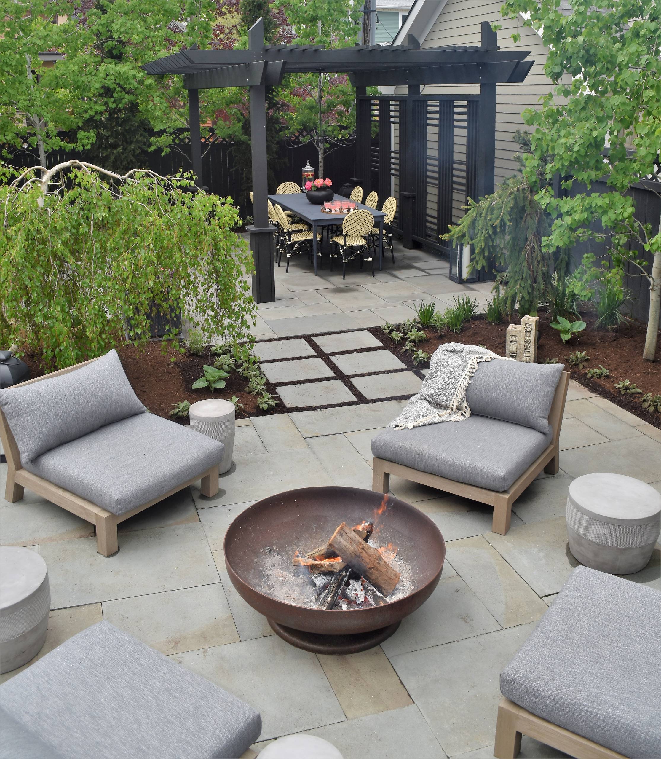 A modern backyard design featuring a cozy seating area with gray cushioned chairs surrounding a fire pit, a dining area under a black pergola, and lush greenery with trees and plants enhancing the ambiance.