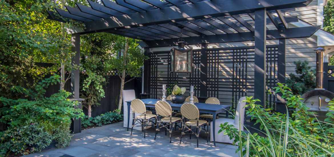 A chic outdoor dining area featuring a black pergola with slatted beams, a rectangular dining table, and elegant wicker chairs. The space is surrounded by lush greenery, a privacy trellis, and a modern pendant light, creating a stylish and secluded ambiance.