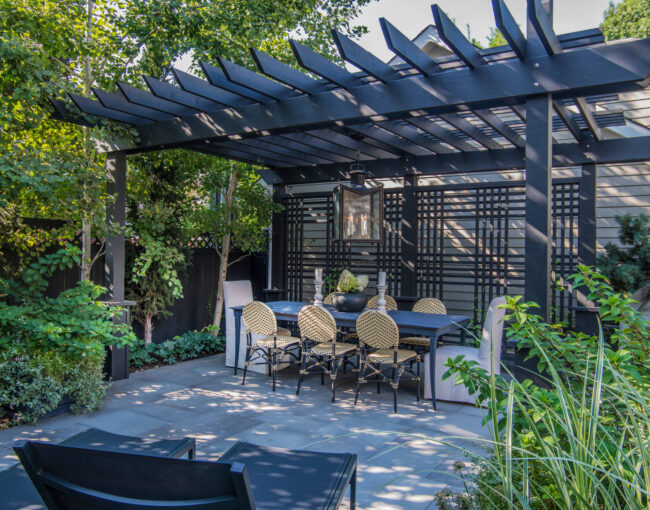 A chic outdoor dining area featuring a black pergola with slatted beams, a rectangular dining table, and elegant wicker chairs. The space is surrounded by lush greenery, a privacy trellis, and a modern pendant light, creating a stylish and secluded ambiance.