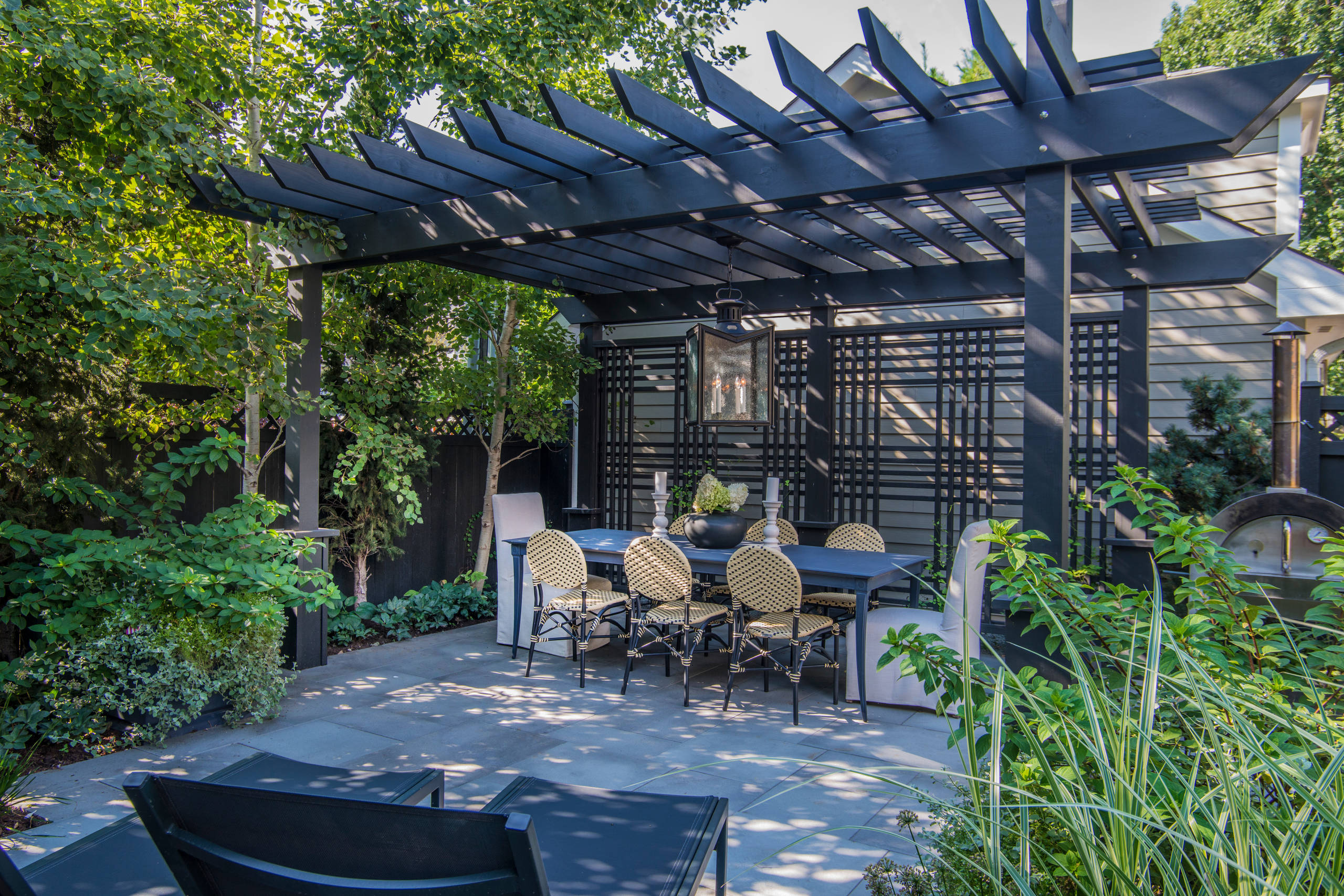 A chic outdoor dining area featuring a black pergola with slatted beams, a rectangular dining table, and elegant wicker chairs. The space is surrounded by lush greenery, a privacy trellis, and a modern pendant light, creating a stylish and secluded ambiance.