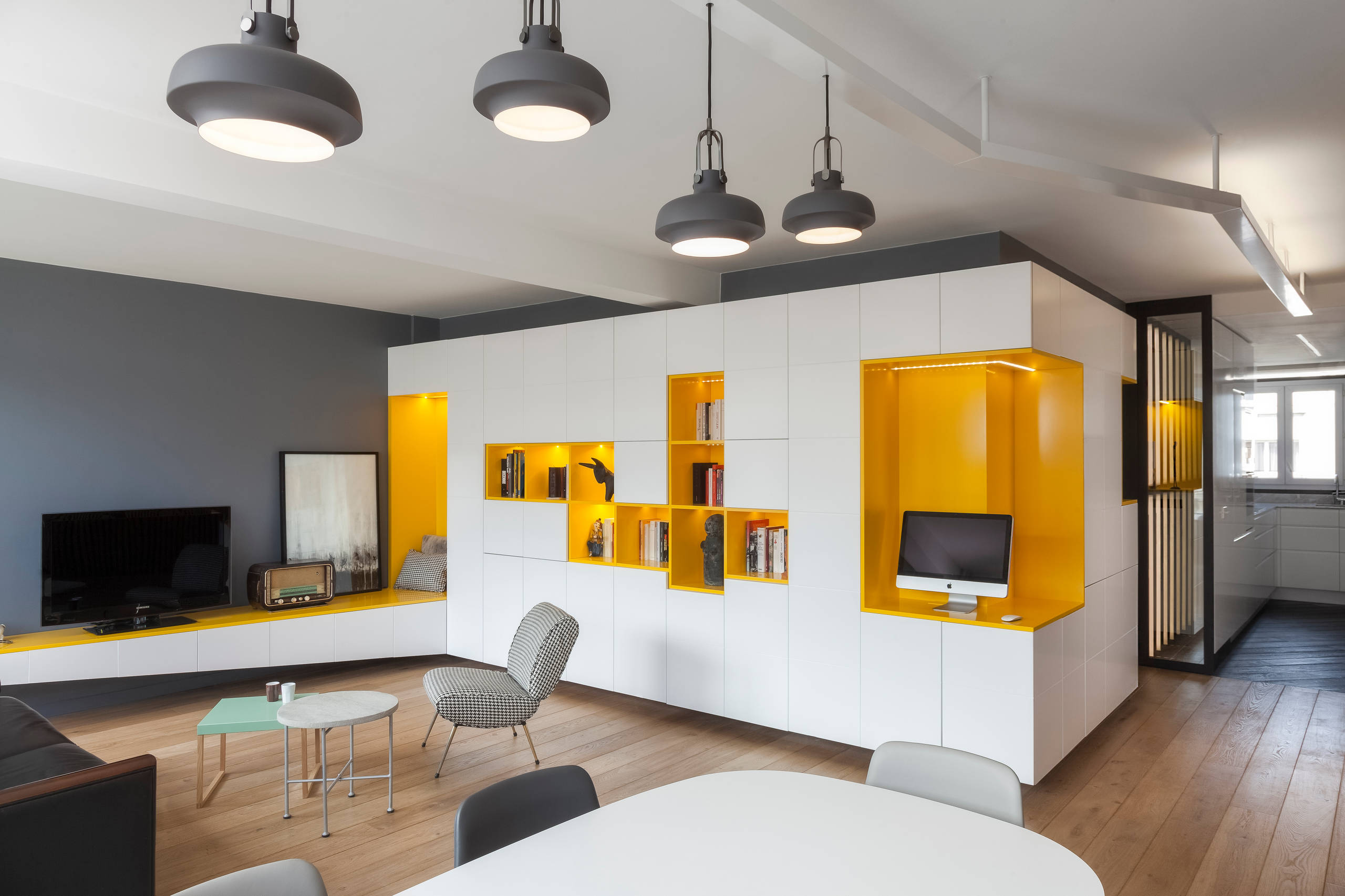 Modern living space with built-in white cabinetry accented by bright yellow shelving, featuring a workspace nook, seating area, and minimalist decor.