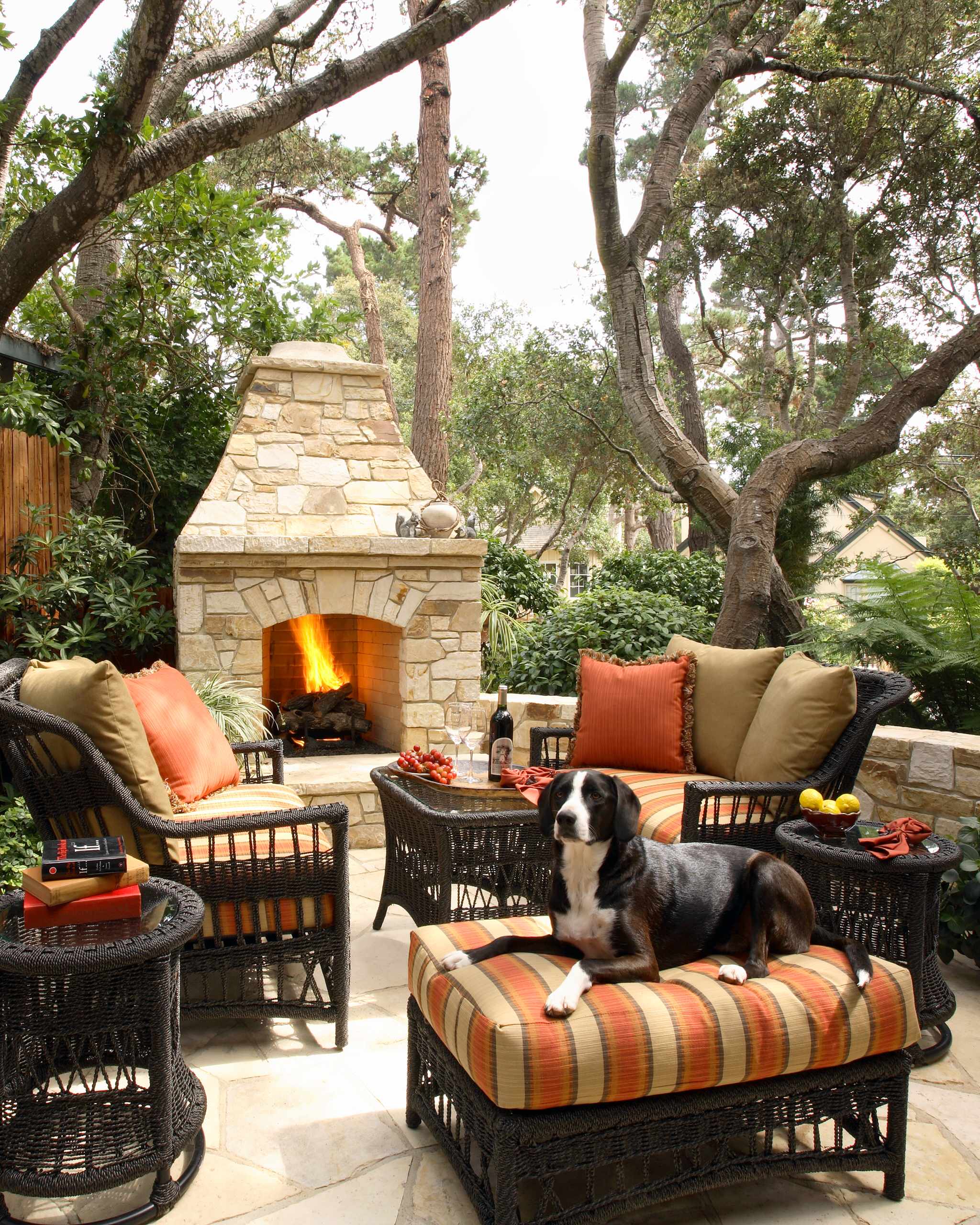 A cozy outdoor seating area with wicker furniture featuring colorful cushions. A dog lounges on a striped ottoman, while a stone fireplace burns brightly in the background among lush greenery and tall trees.