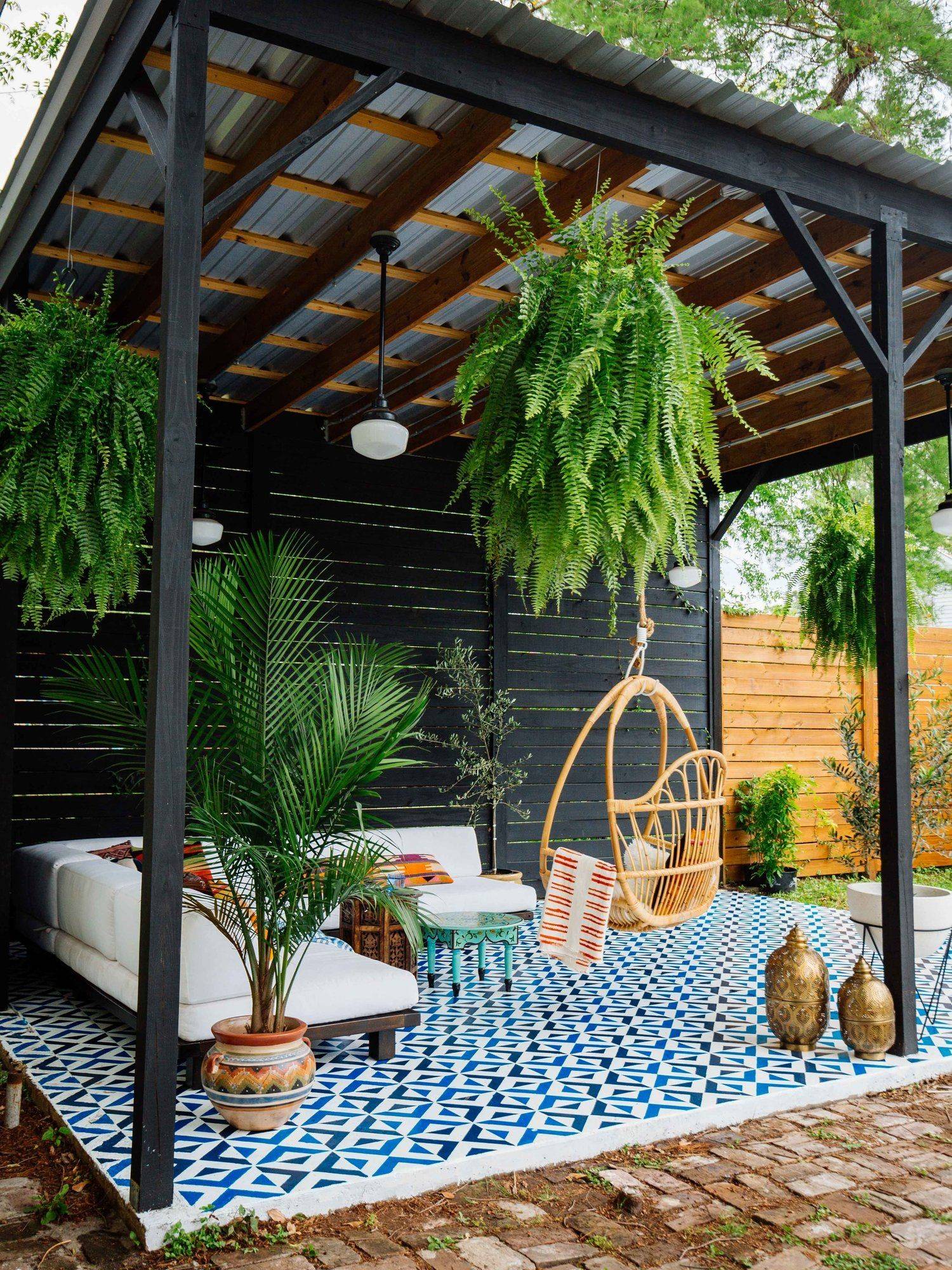 A vibrant patio featuring a geometric blue-and-white tiled floor, a cozy white sectional sofa, a hanging rattan chair, and lush greenery, including hanging ferns and potted plants. The space is enclosed by black and wooden privacy walls under a metal roof.