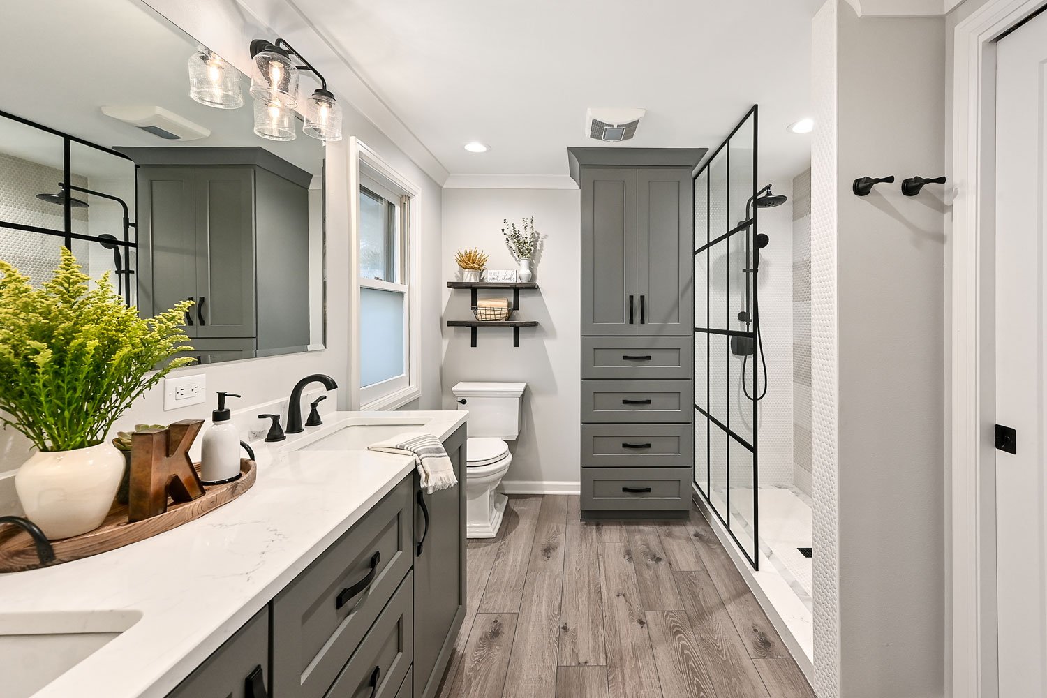 Modern bathroom with gray cabinetry, black fixtures, glass shower enclosure, and wood-look flooring.