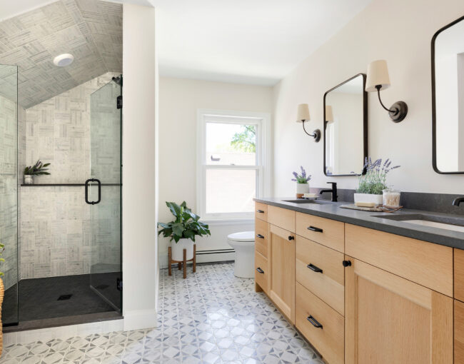 Bright bathroom with wood double vanity, glass shower, and patterned tile floor.