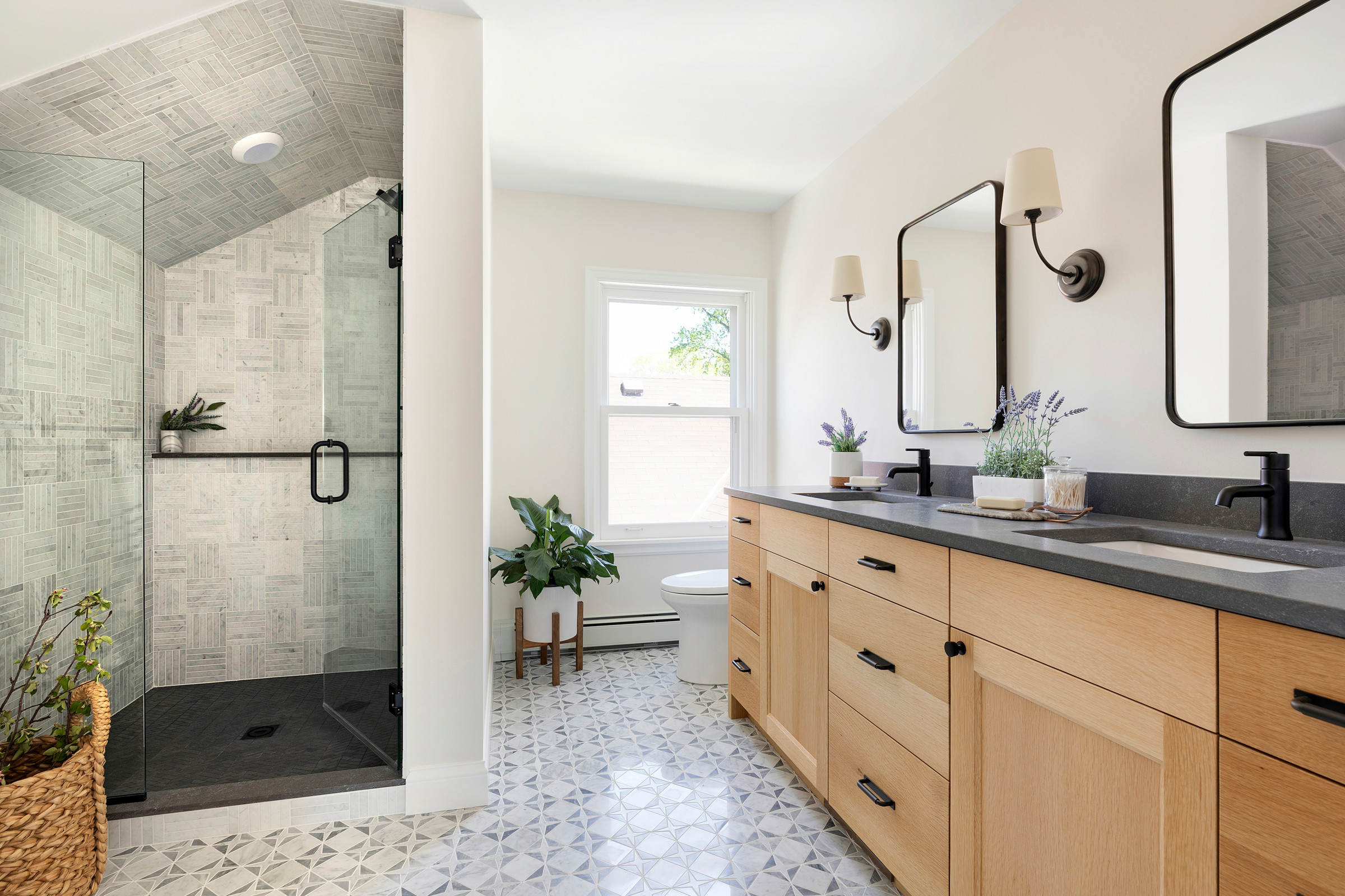 Bright bathroom with wood double vanity, glass shower, and patterned tile floor.