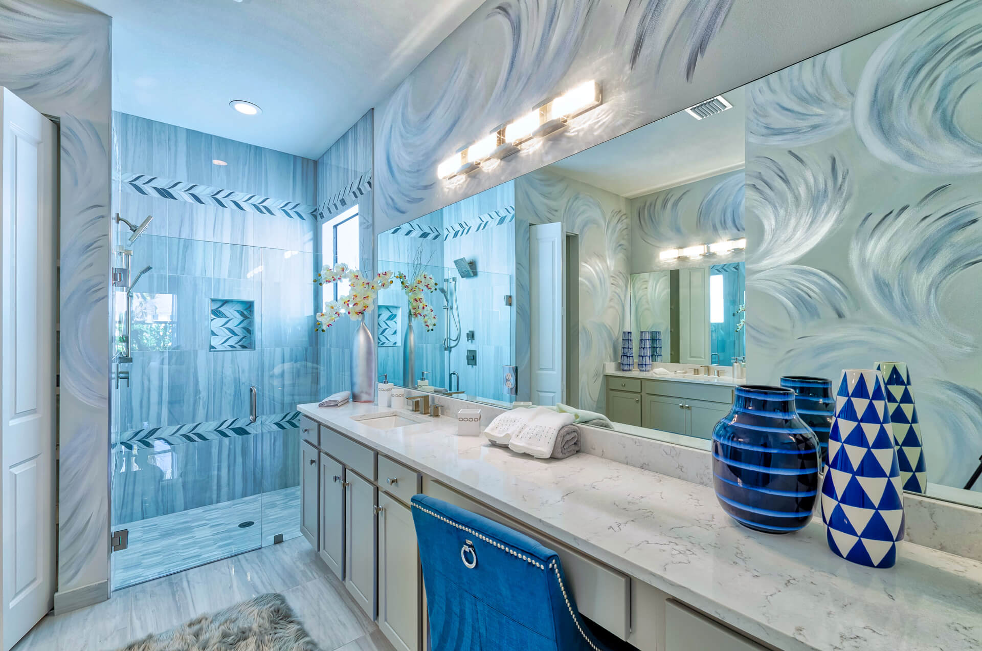 Luxurious modern bathroom featuring a long marble-top vanity with dual sinks, sleek cabinetry, and elegant silver fixtures. A glass-enclosed shower with blue and gray patterned tile walls complements the swirled artistic wall design. Decorative blue ceramic vases and a plush blue chair add vibrant accents, while large mirrors enhance the space's brightness.