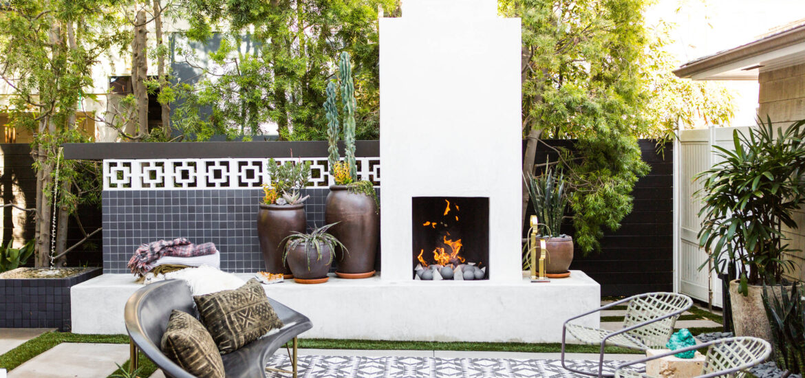 A stylish outdoor space featuring a black and white geometric rug, a modern black sofa with patterned pillows, and a clean, white outdoor fireplace. The area is surrounded by potted plants and trees, creating a serene atmosphere for relaxation.