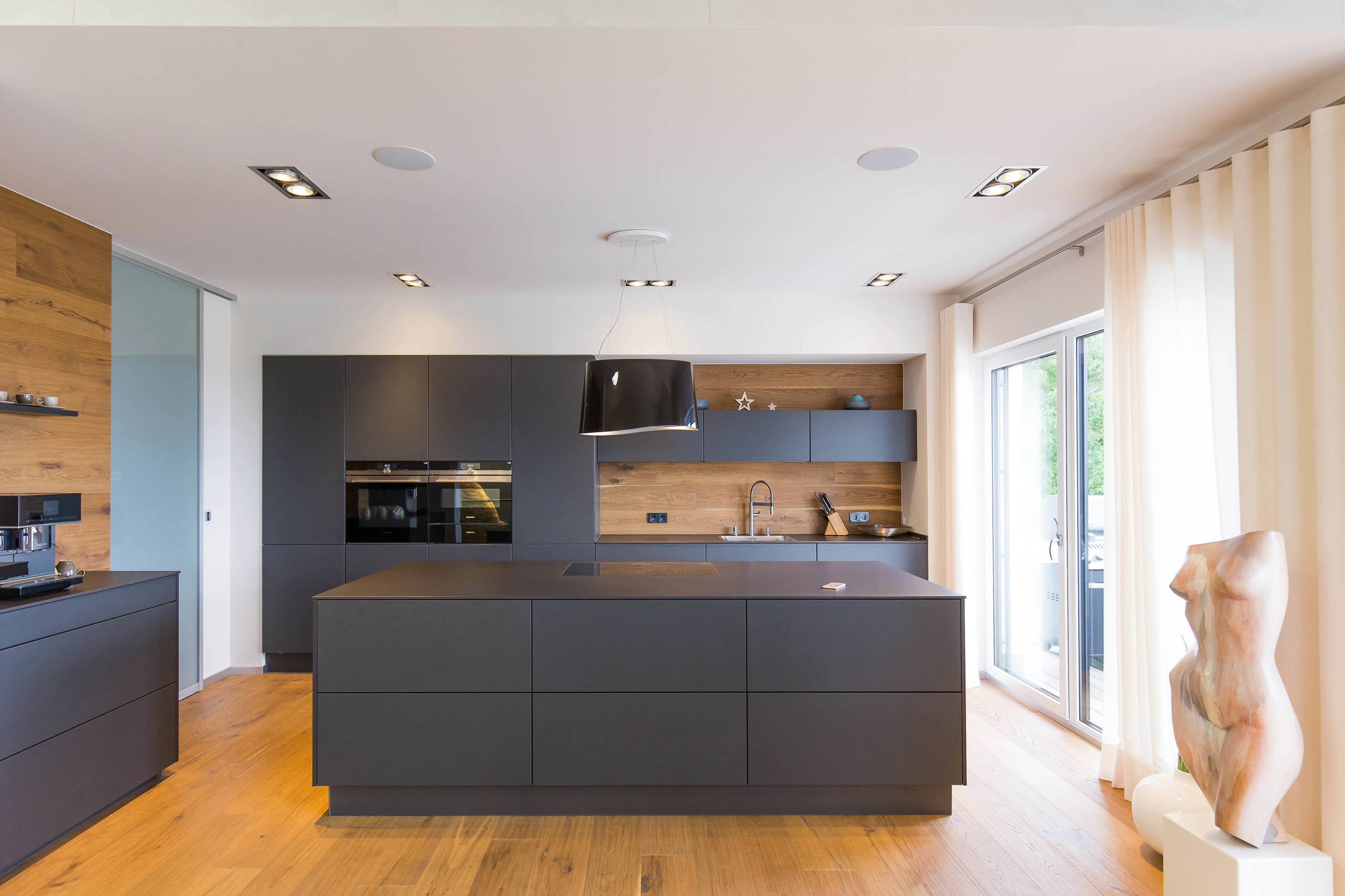 Minimalist modern kitchen with matte black cabinets, wood backsplash, and matching wood flooring.