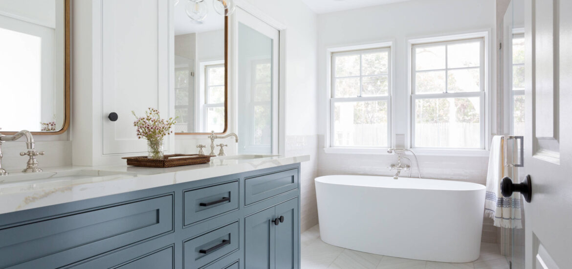 Bright bathroom with blue vanity, white freestanding bathtub, and large windows.