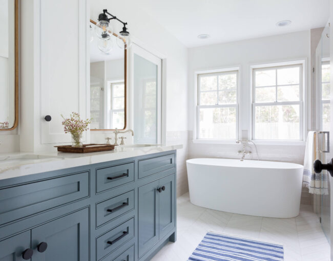 Bright bathroom with blue vanity, white freestanding bathtub, and large windows.