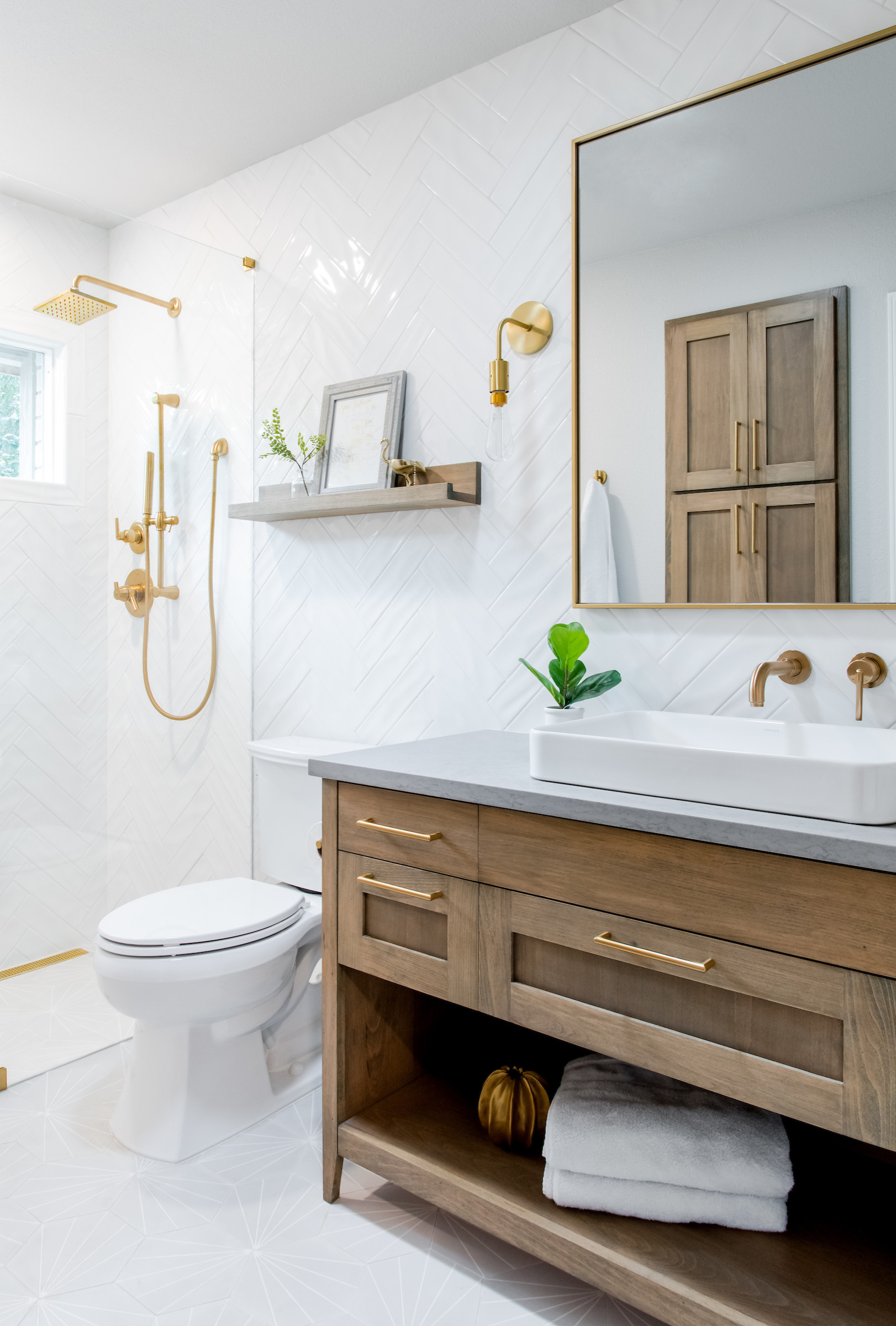 Modern farmhouse bathroom with gold fixtures, wooden vanity, and white herringbone tile walls.