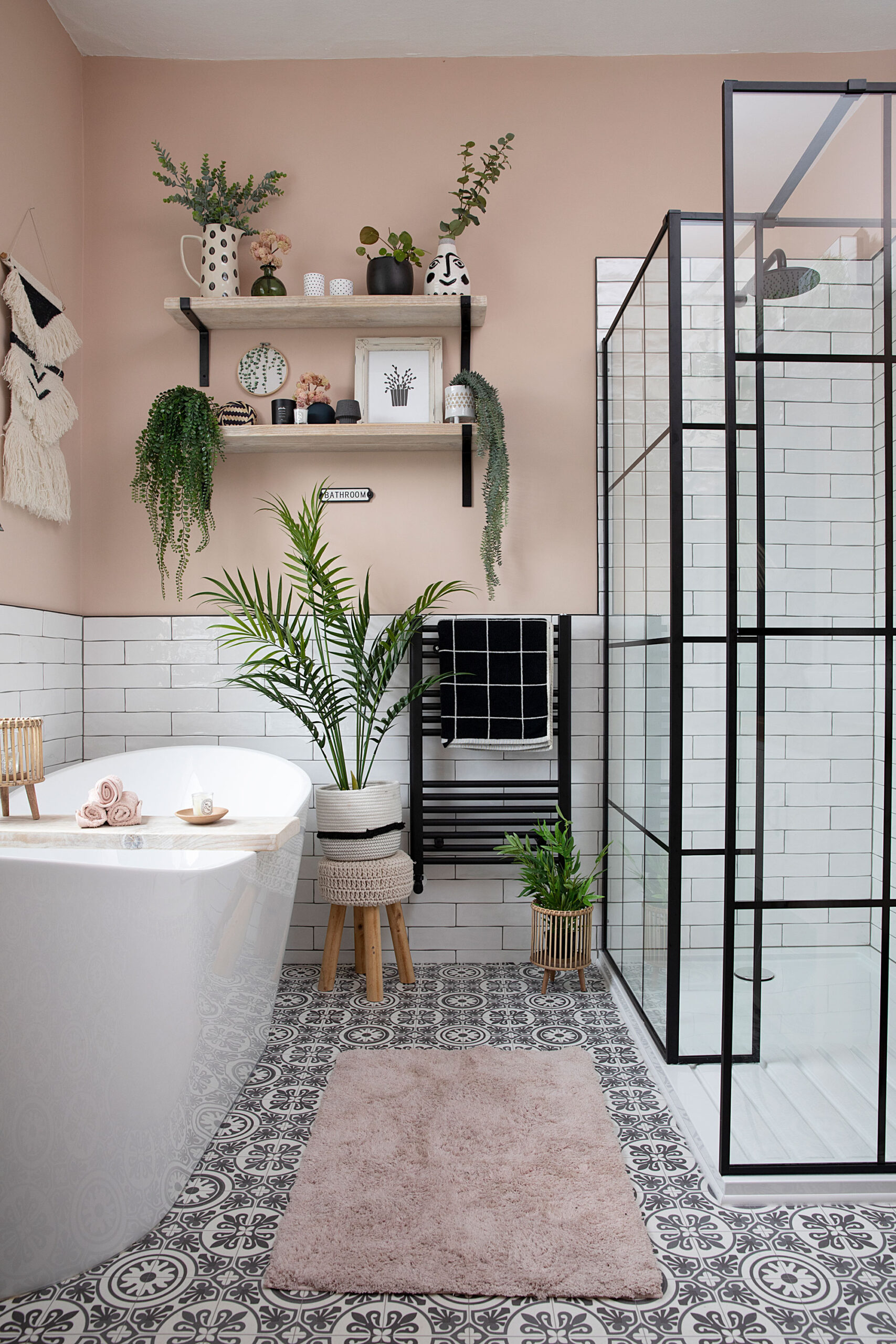 Chic bohemian bathroom with a freestanding bathtub, patterned floor tiles, white subway walls, black-framed glass shower, pink walls, and decorative plants.