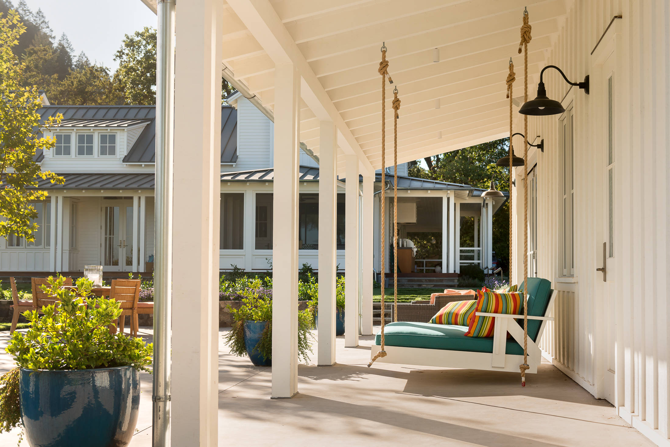 A bright and airy farmhouse-style porch featuring a turquoise swing with colorful striped cushions, hanging by thick ropes from a white-beamed ceiling. Large blue ceramic planters with lush greenery frame the space, while the view extends to a landscaped yard and a modern farmhouse with metal roofing.