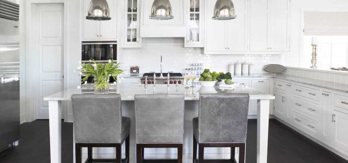 Bright white kitchen with marble island, stainless steel accents, and gray upholstered bar stools.