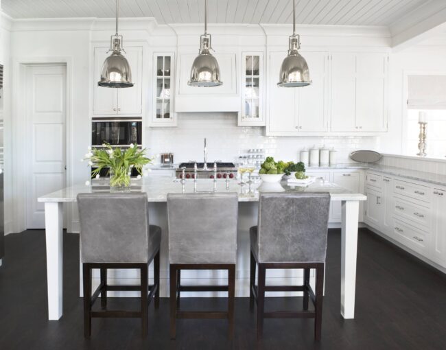 Bright white kitchen with marble island, stainless steel accents, and gray upholstered bar stools.