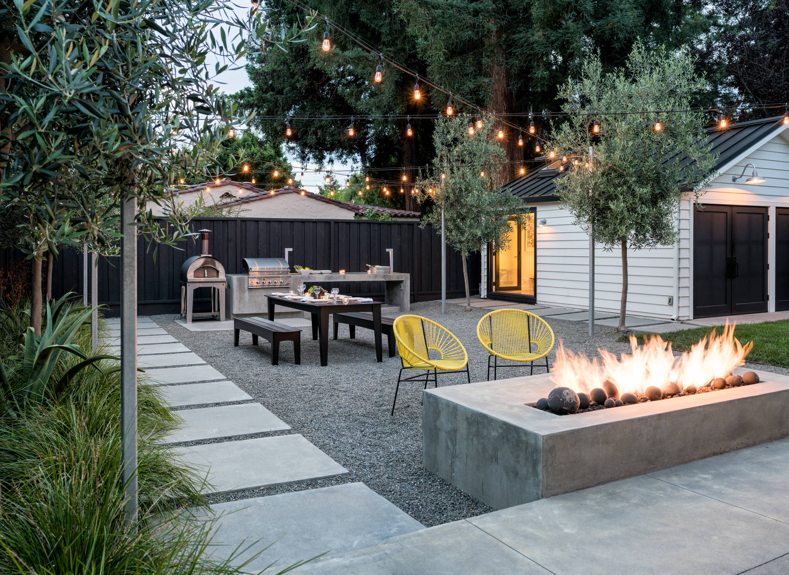 A modern backyard design featuring a fire pit surrounded by concrete seating, string lights overhead, a dining table with benches, and a pizza oven on the left. The area is landscaped with trees and greenery, complemented by a sleek black fence and white building in the background.