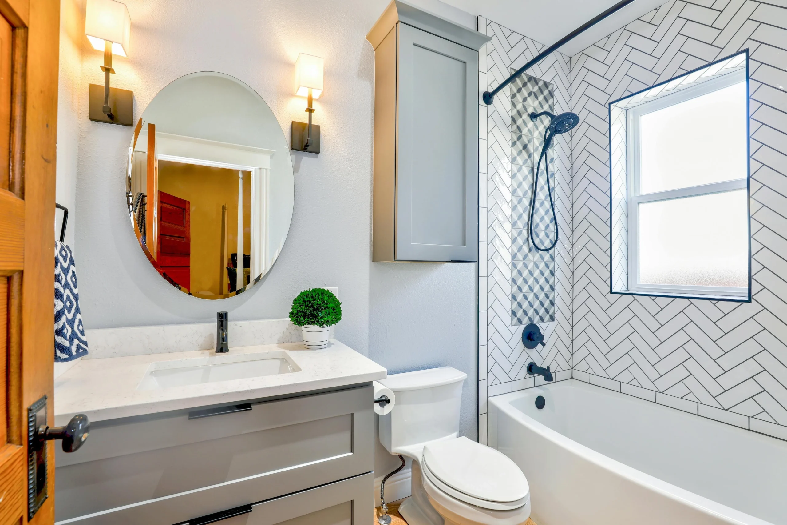 Small modern bathroom with herringbone tile bathtub surround, gray vanity, and round mirror.