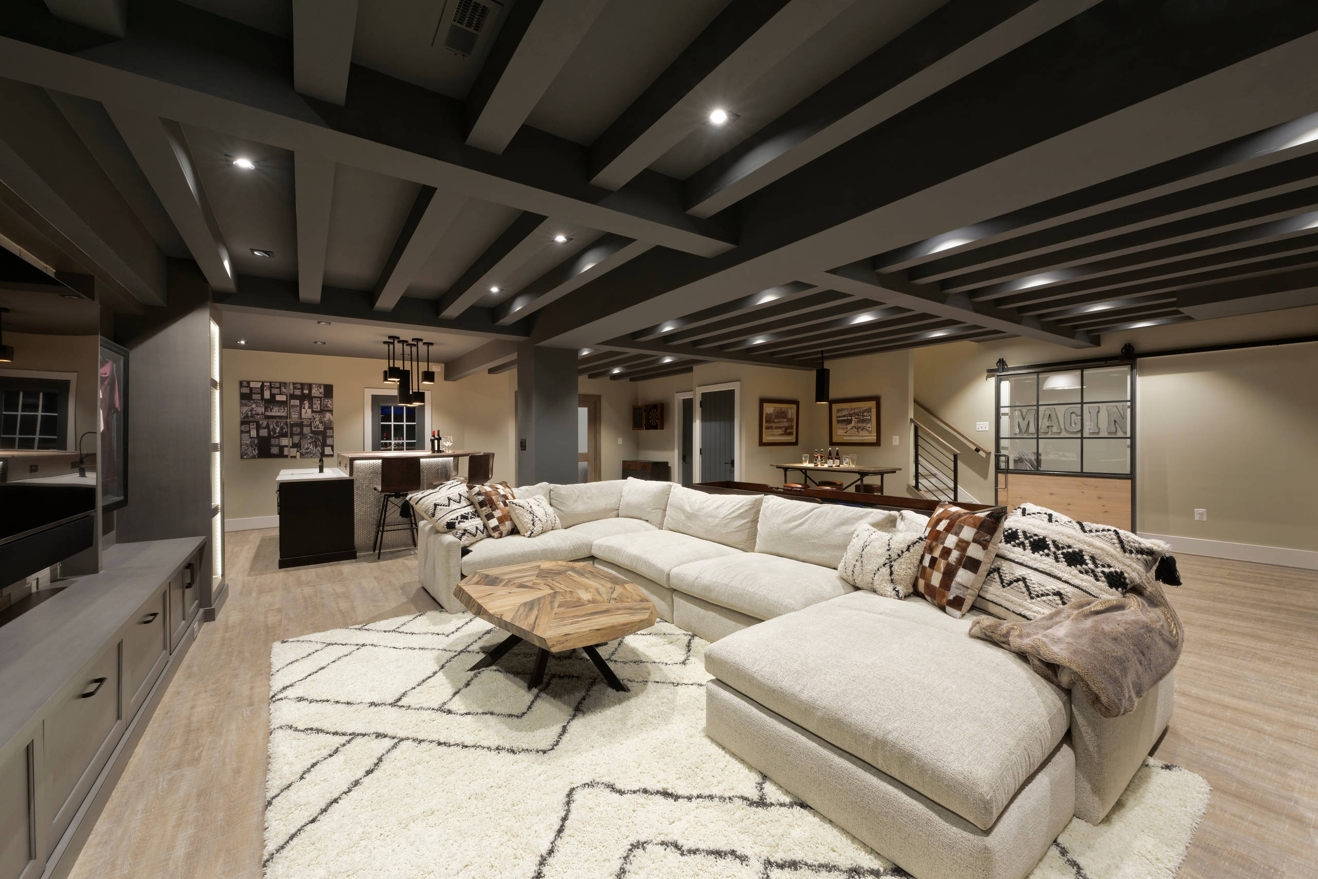 Modern basement with sectional sofa, bar area, and decorative ceiling beams.