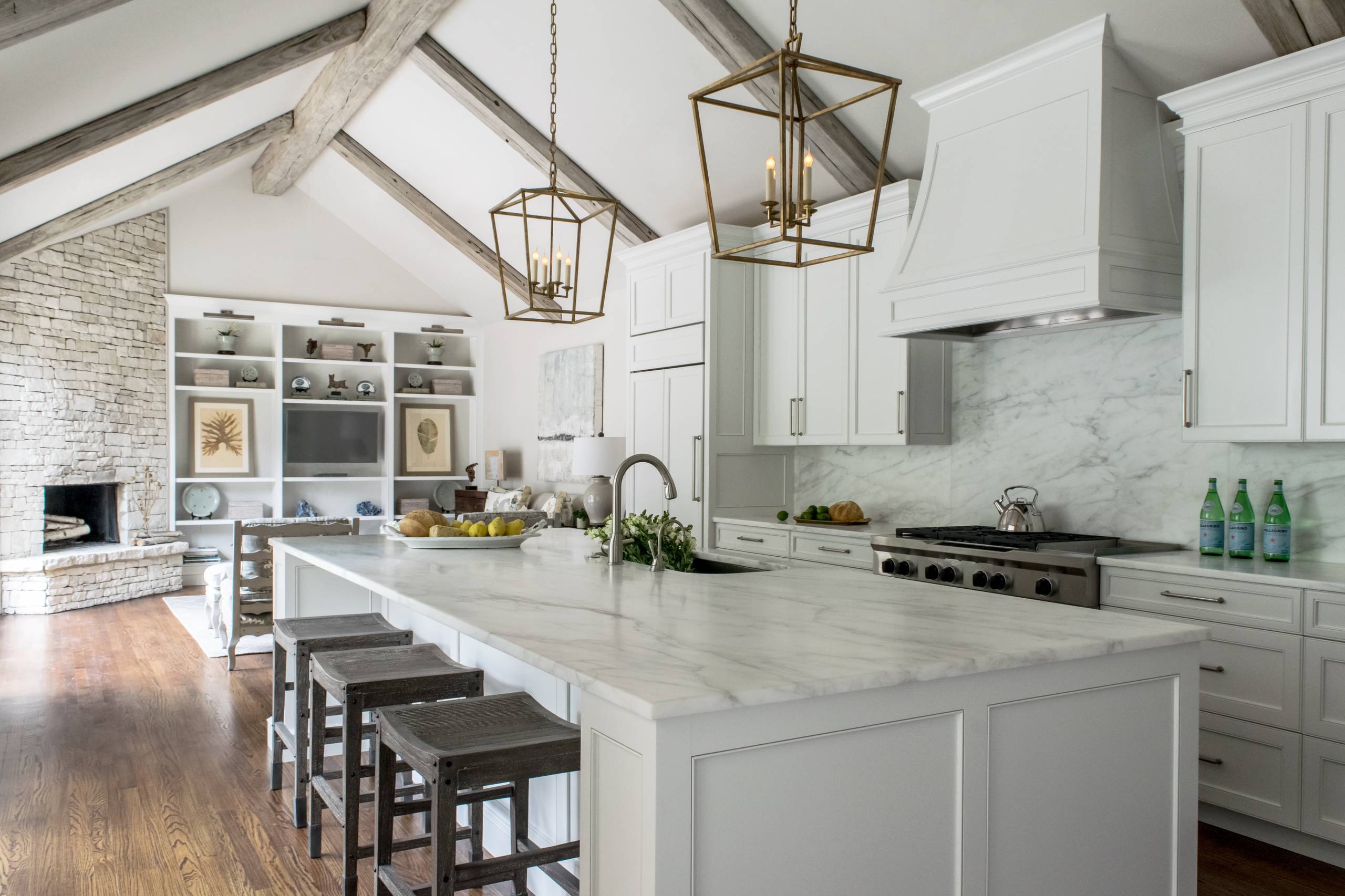 Bright kitchen with a large marble island, white cabinetry, exposed wood beam ceiling, and stone fireplace in the background.