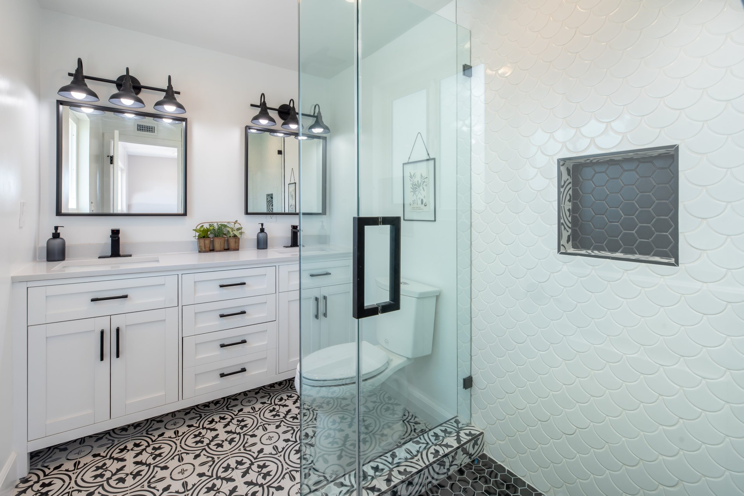 Bright modern bathroom with white cabinetry, black hardware, patterned tile flooring, and a glass shower with fish scale wall tiles and hexagon niche.