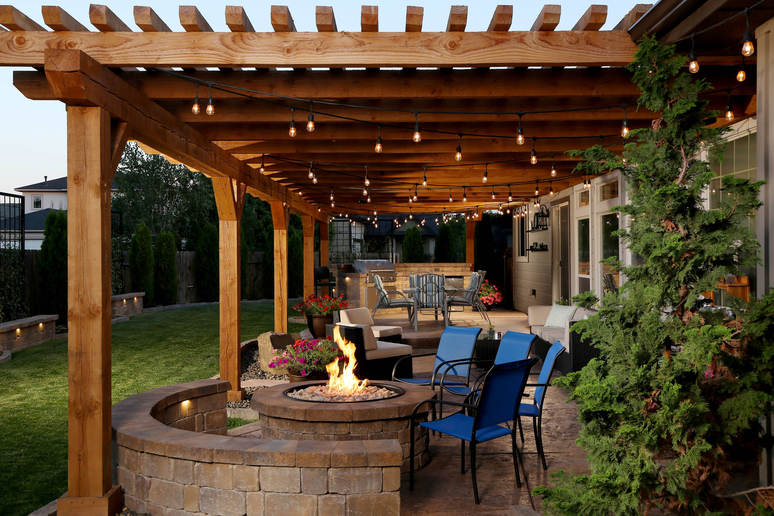 A wooden pergola with string lights over a cozy outdoor seating area, featuring a fire pit surrounded by stone benches and colorful plants.