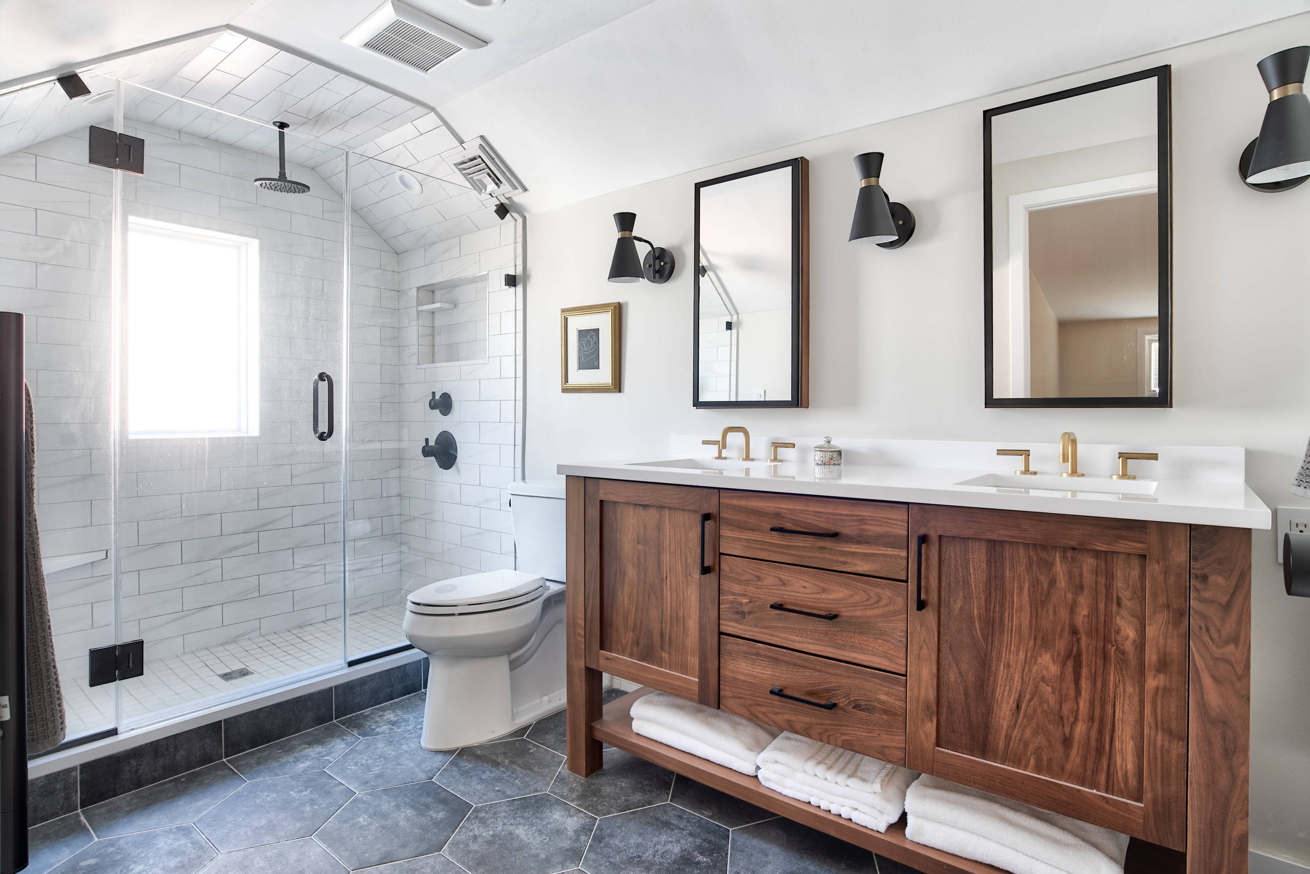Modern bathroom featuring a glass-enclosed shower with white subway tiles, dual rainfall showerheads, and a wooden vanity with white countertop, brass faucets, and open shelf storage beneath.