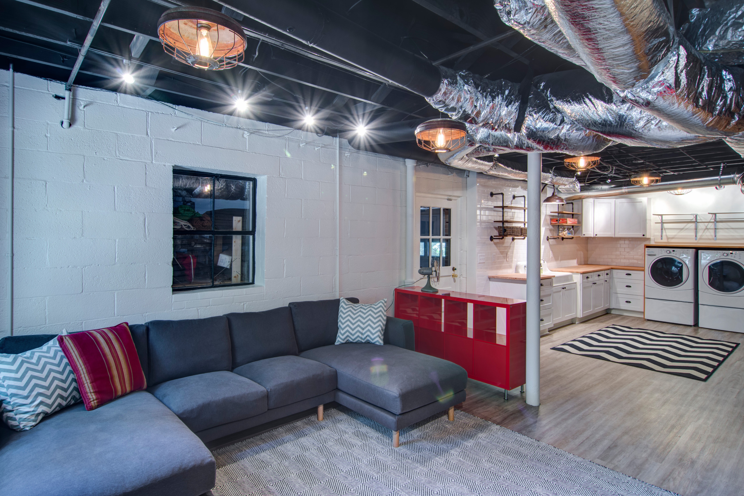 Modern finished basement with gray sectional sofa, laundry area, and open ceiling design.
